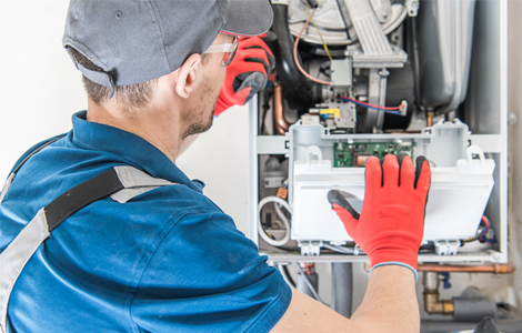 A technician kneeling at a gas furnace in a basement during the annual inspection, the visit that checks for furnace carbon monoxide before winter.